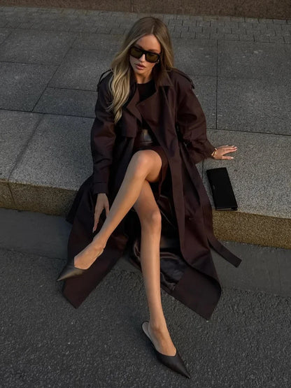 Woman in a long dark coat sitting on a street corner wearing sunglasses and high heels.