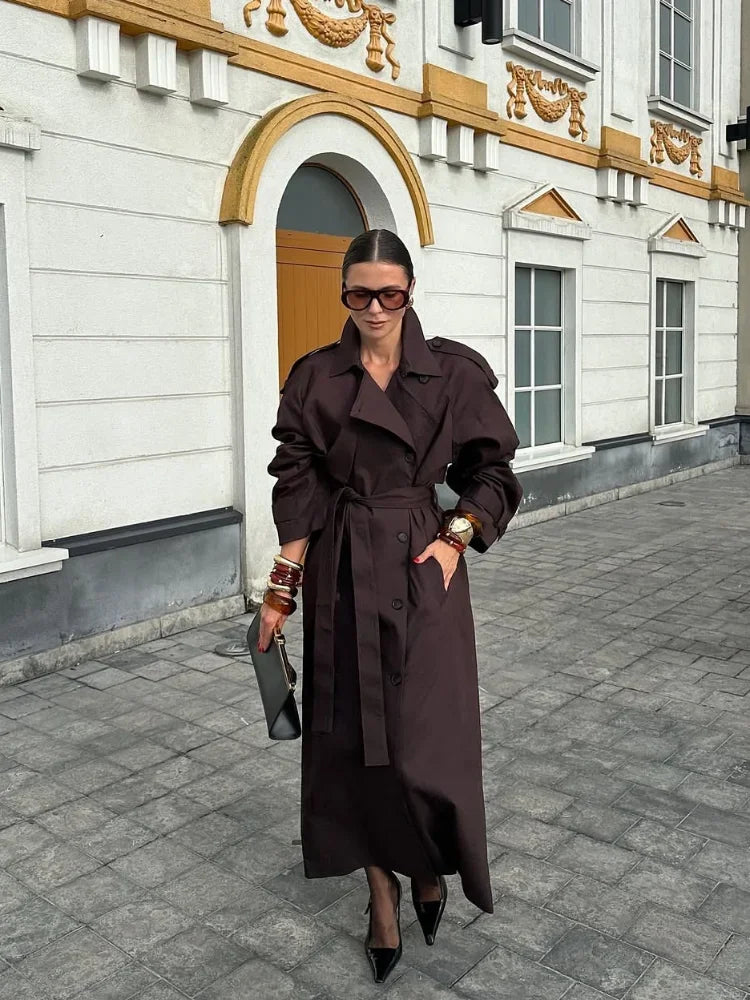 Woman in a long brown coat standing on a street with decorative building facade.