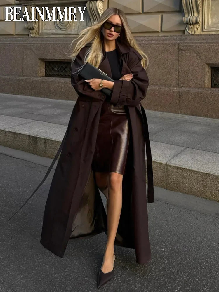 Woman in a long brown coat and sunglasses standing on a city street.
