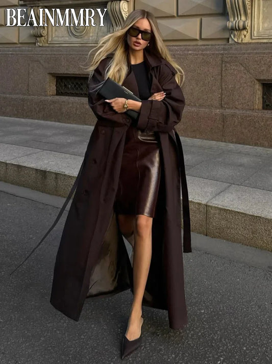 Woman in a long brown coat and sunglasses standing on a city street.