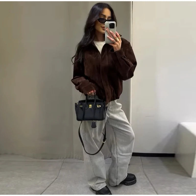 Person taking a mirror selfie wearing a brown jacket, white pants, and holding a black handbag.