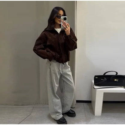 Person taking a mirror selfie wearing a brown jacket and light-colored pants in a minimalistic room.