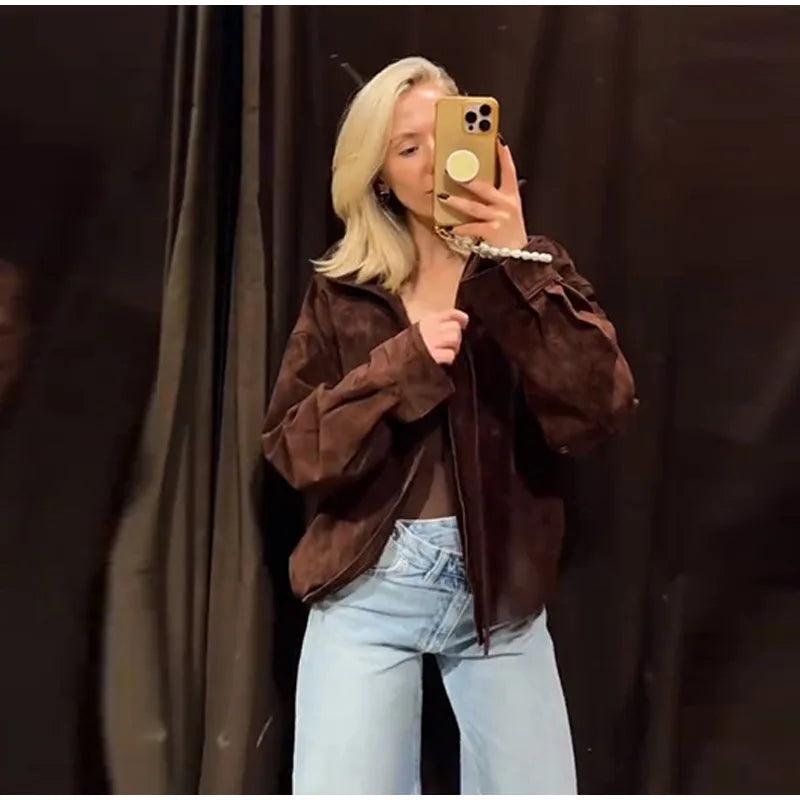 Person wearing a brown jacket and light blue jeans taking a mirror selfie against a dark curtain.
