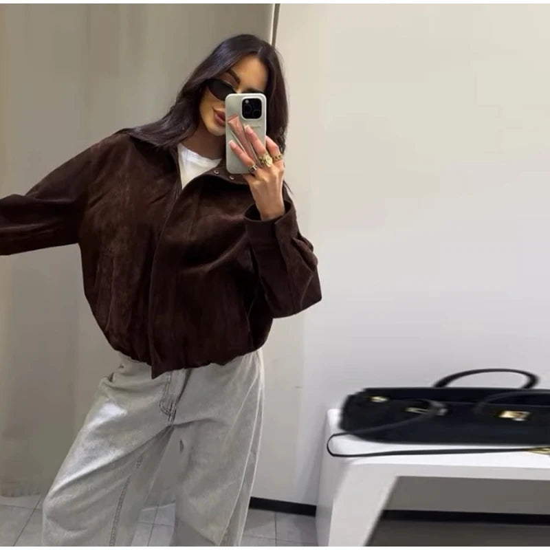 Person taking a mirror selfie wearing a brown jacket and light-colored pants, with a black handbag on a white surface in the background.
