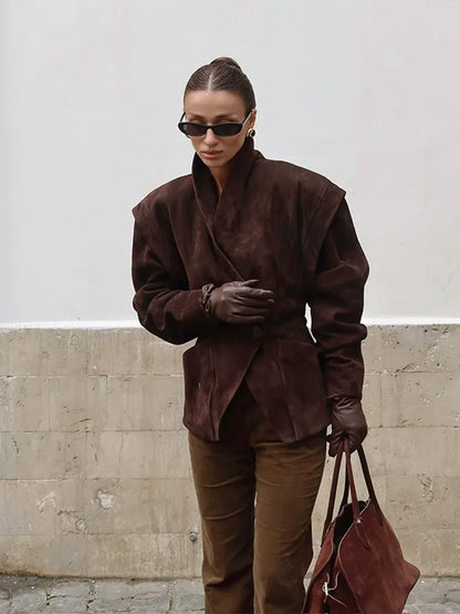 Person wearing a brown suede outfit with sunglasses and holding a matching bag against a white wall.
